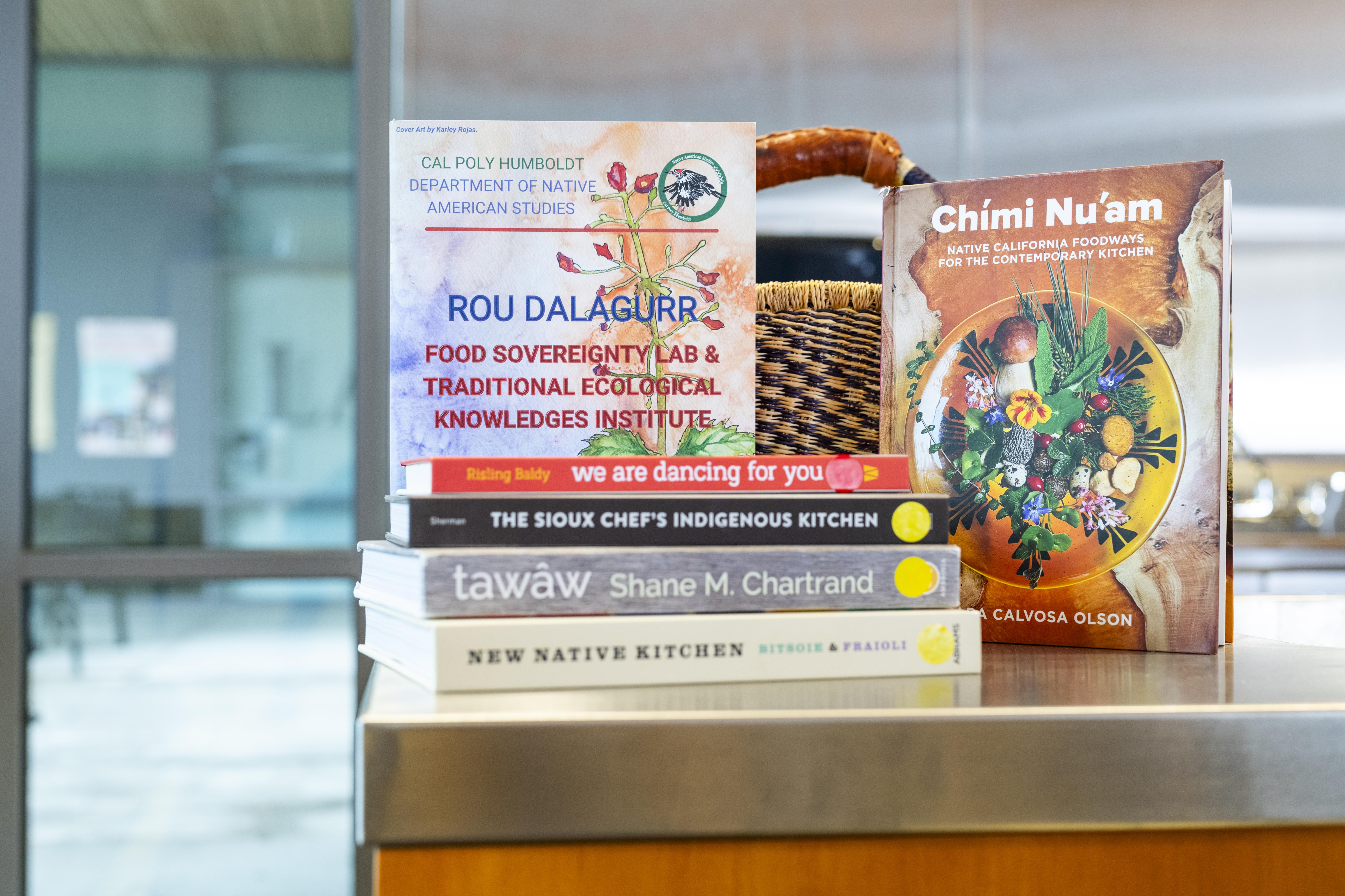 A stack of Indigneous Cookbooks and one of the FSL Reports, a woven basket is in the background